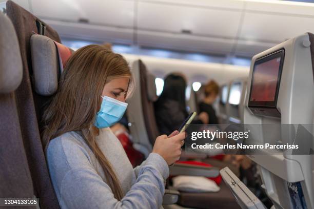 profile of a young woman in a mask using the mobile while sitting inside an airplane - classe virtuelle stock-fotos und bilder