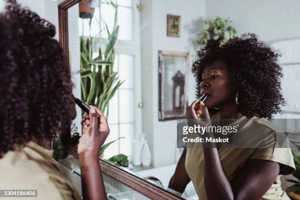 mature afro female beautician applying lipstick while looking at mirror reflection in bathroom - lipstick stock pictures, royalty-free photos & images