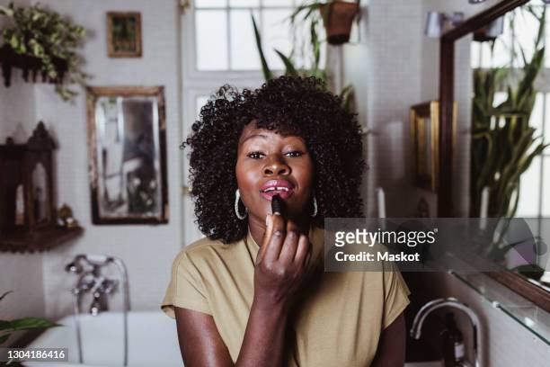 portrait of mature afro female make-up artist applying lipstick in bathroom at home - bad news stock-fotos und bilder