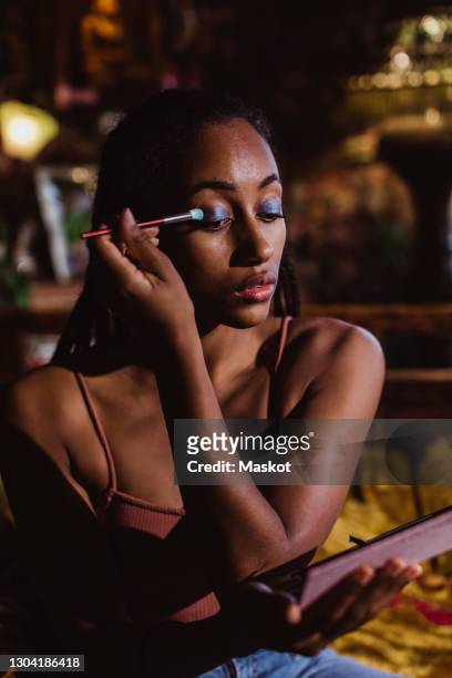 young woman applying eyeshadow while sitting in bedroom at home - applying makeup with brush fotografías e imágenes de stock