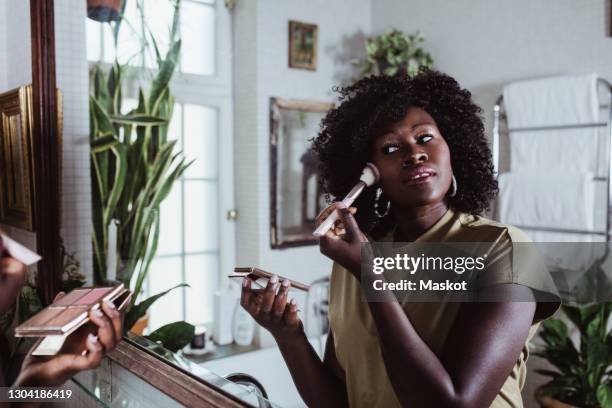 reflection of mature afro female influencer applying foundation make-up in bathroom at home - sminkborste bildbanksfoton och bilder