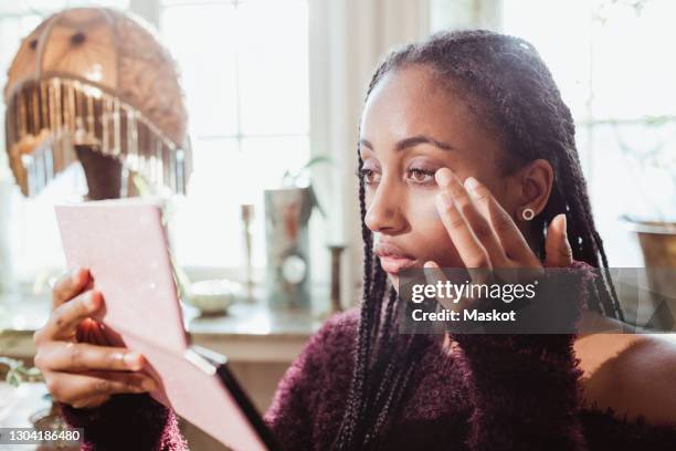 young woman looking at hand mirror while applying make-up at home - make up looks stock pictures, royalty-free photos & images