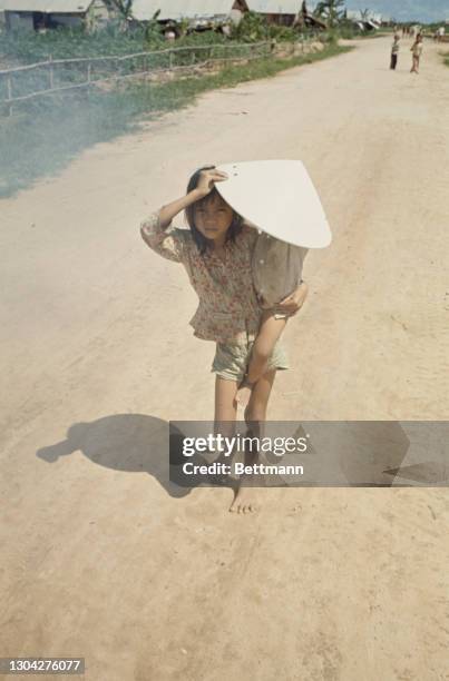 Young Vietnamese girl carrying a smaller child in her arm, sheltering them both from the sun with a traditional non la hat, among the displaced...