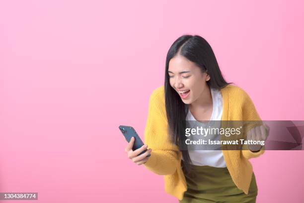 cheerful young asian woman using smartphone and receiving good news from the message on mobile chat application isolated over pink background - euphoric female stock pictures, royalty-free photos & images