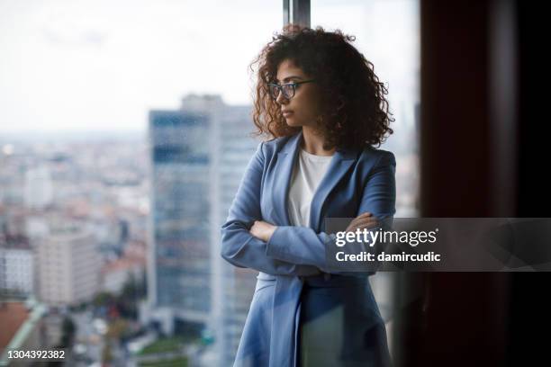 jonge nadenkende onderneemster die een kostuum draagt dat zich in haar bureau bevindt en door venster kijkt - stress management stockfoto's en -beelden