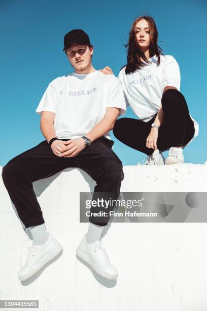 fashionable young couple sitting on concrete wall modern fashion portrait - tee stock pictures, royalty-free photos & images