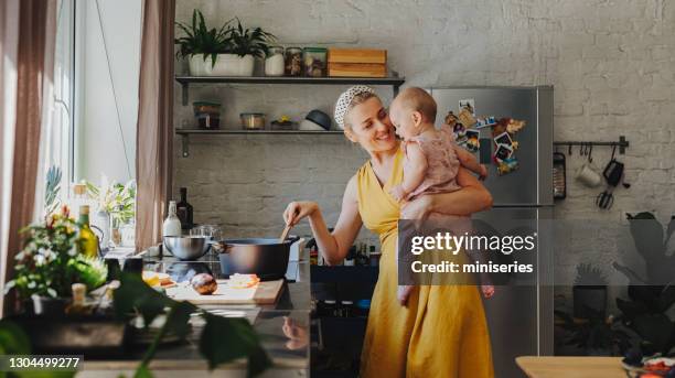 a young smiling caucasian mother holding her baby girl while preparing lunch at home - yellow kitchen stock pictures, royalty-free photos & images