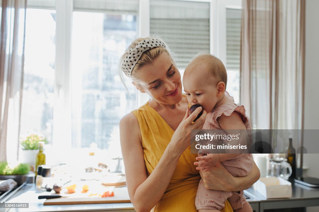 An Adorable Baby Girl Biting a Plum while Being Held by Her Mother in a Kitchen at Home