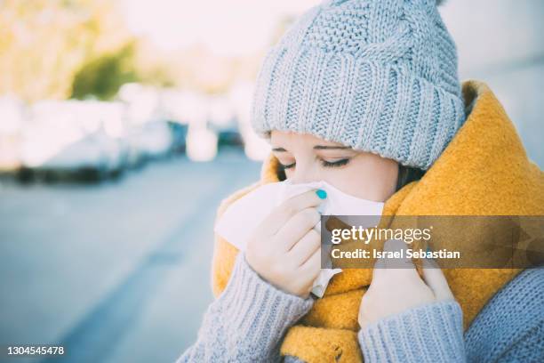 close up view of a young caucasian woman dressed in winter clothing as she blows her nose from being ill. - winter illness stock pictures, royalty-free photos & images