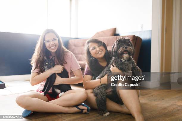 portrait of two smiling young women sitting on living room floor holding a cat and a dog - cat and owner stock pictures, royalty-free photos & images