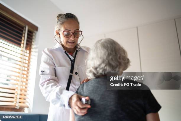 自宅��で上級女性を試験するために聴診器を使用する医師 - listening to heartbeat ストックフォトと画像