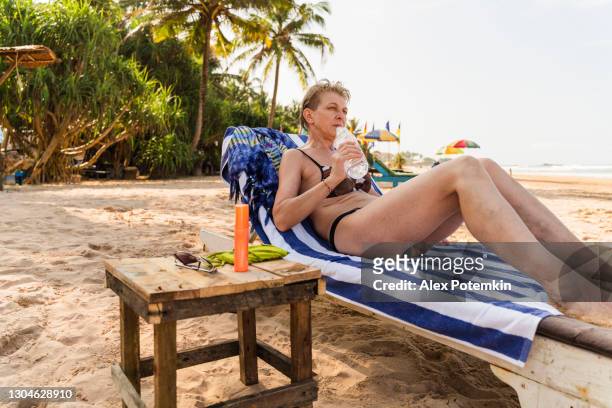un'attraente donna europea attiva di 50 anni, una turista, riposa in una chaise lounge su una spiaggia di bentota, nello sri lanka. sta bevendo acqua da una bottiglia di plastica. - 50 54 years foto e immagini stock