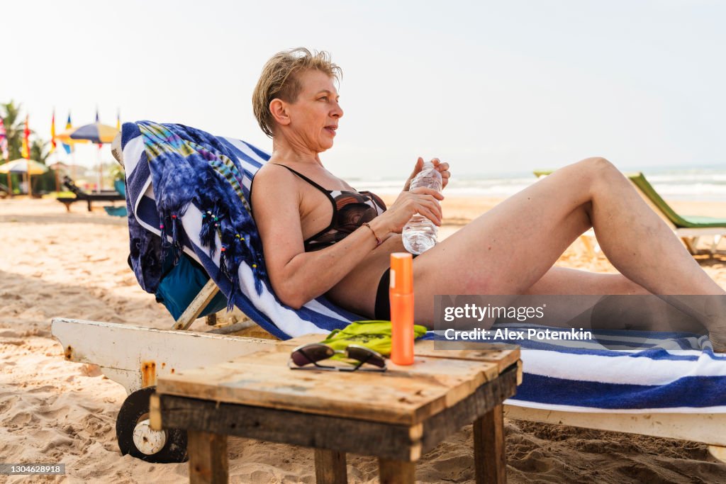 Una atractiva mujer europea de 50 años, una turista, descansa en un salón chaise en una playa de Bentota, Sri Lanka. Está bebiendo agua de una botella de plástico.