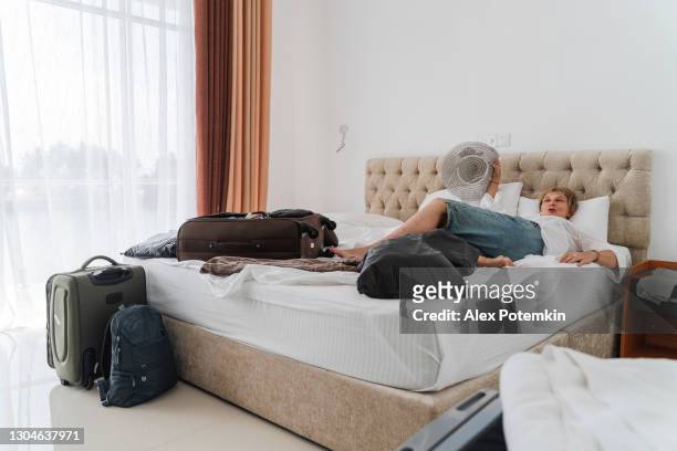 aantrekkelijke kortharige kaukasische 50-jarige vrouw, een toerist, ligt en rust op een bed naast een gedeeltelijk uitgepakte koffer in de moderne lichte hotelkamer, moe van het inpakken van spullen. - 50 54 years stockfoto's en -beelden