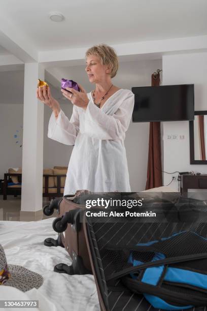 atractiva mujer caucásica de pelo corto de 50 años, una turista, está empacando una maleta en la habitación contemporánea del hotel brillante preparándose para un viaje futuro, sosteniendo regalos envueltos recientemente comprados. - 50 54 years fotografías e imágenes de stock