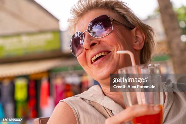 het portret van de close-up van de gelukkige lachende europese 50-jaar-oude toeristenvrouw in het openluchtrestaurant op het strand, dat haar mangococktail met een het drinken stro drinkt - 50 54 years stockfoto's en -beelden