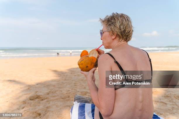 attractive mature 50-years-old woman, the european tourist, enjoying the fresh coconut water on a tropical beach. - alex potemkin coronavirus stock pictures, royalty-free photos & images