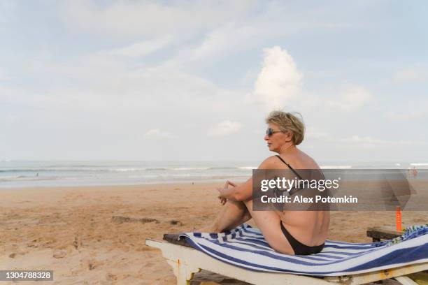 clouds don't prevent tanning and sunbathing in the south asian tropical climate. a mature attractive 50-years-old european woman, the tourist, is sitting on a beach bed in sri lanka. - alex potemkin coronavirus stock pictures, royalty-free photos & images