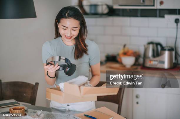 mulher asiática chinesa bonita recebendo desempacotamento experimentando em seu fone de ouvido de compras on-line na cozinha - unboxing - fotografias e filmes do acervo