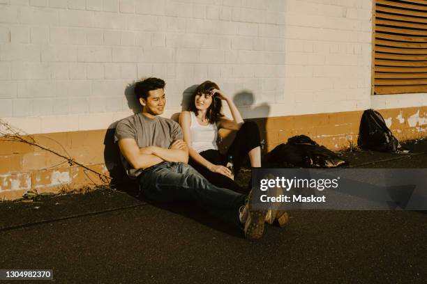 male and female friends sitting side by side against wall on footpath - fianco a fianco foto e immagini stock