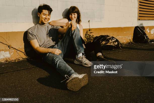 smiling man sitting with female friend on footpath against wall - beste freunde teenager stock-fotos und bilder