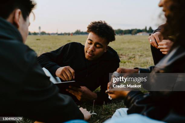 young man holding book sitting with friends in park - small group sitting in grass photos et images de collection