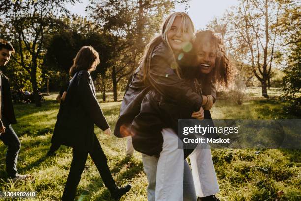 young woman carrying piggyback to female friend in park - piggyback stock pictures, royalty-free photos & images