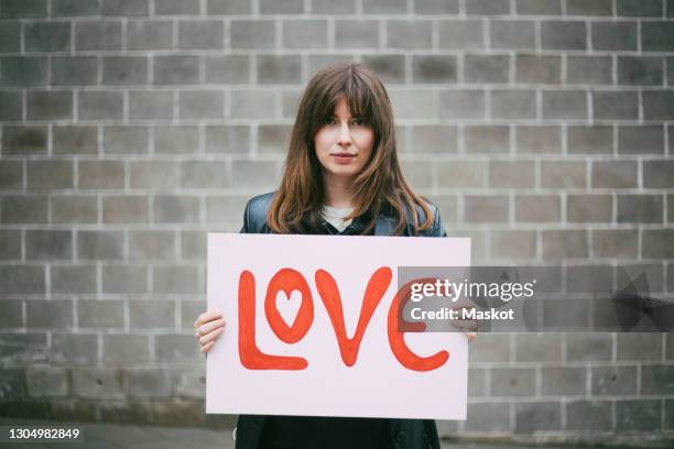 portrait of female activist with love poster against wall - frau mit plakat stock-fotos und bilder