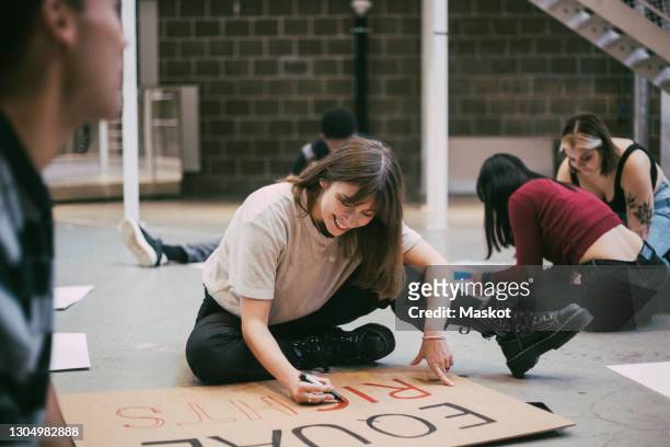 smiling woman preparing signboard while sitting with male and female activists in building - giustizia sociale foto e immagini stock