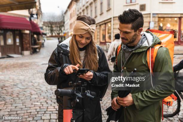 heterosexual couple using smart phone while standing electric push scooter on street in city - bike sharing stock pictures, royalty-free photos & images