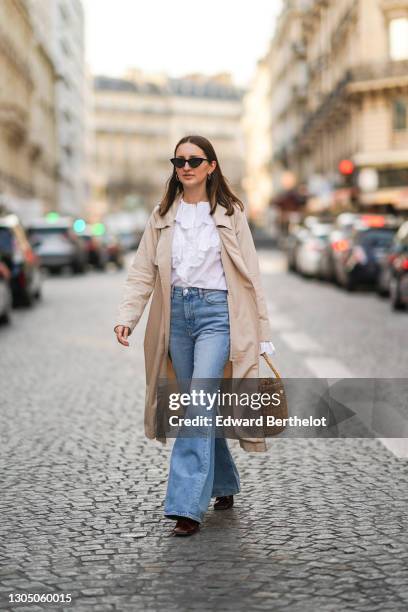 Marielle Haon wears sunglasses, a white ruffle shirt, a beige trench long coat, blue denim flare jeans pants, a brown wicker basket bag, burgundy...