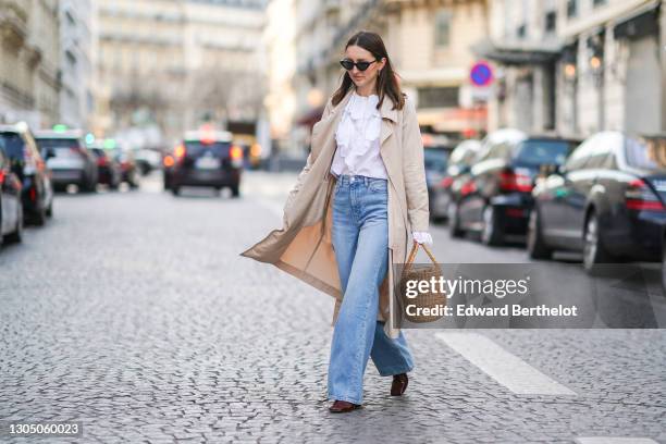 Marielle Haon wears sunglasses, a white ruffle shirt, a beige trench long coat, blue denim flare jeans pants, a brown wicker basket bag, burgundy...