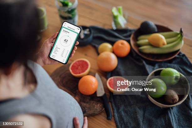 pregnant woman using mobile app to track nutrition and count calories with smartphone while making fresh fruit juice - juice extractor stock pictures, royalty-free photos & images