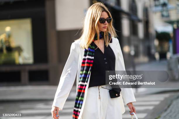 Natalia Verza aka Mascarada wears sunglasses, earrings, a long colored striped scarf, a black vest, a white oversized blazer jacket, a small Chanel...