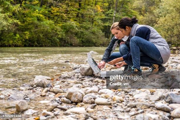 low angle view of female biological researchers squatting at riverside and discussing measured data. - environmentalist stock pictures, royalty-free photos & images