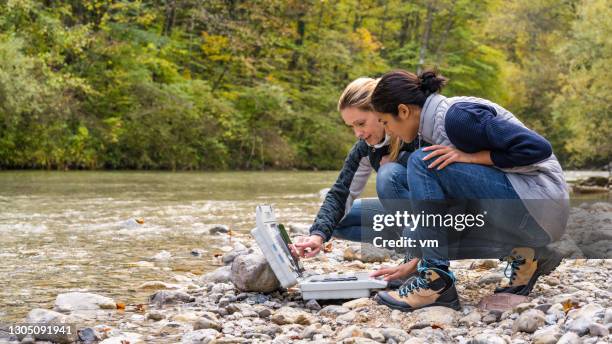 female colleagues discussing data in digital tablet. two women squatting at riverside. - environmentalist stock pictures, royalty-free photos & images