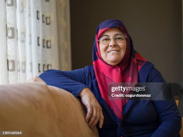 muslim senior woman wearing a headscarf sitting on a sofa at home, daylight portrait - turkish woman stock pictures, royalty-free photos & images