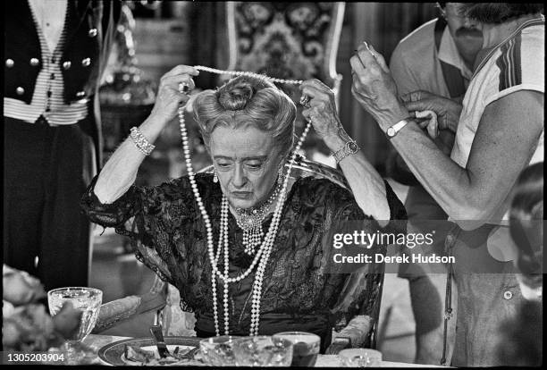 American actress Bette Davis dressed in a black lace dress with strings of pearls hanging down to her waist, an ornate pearl necklace around her neck...