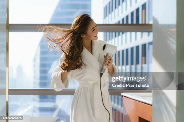 asian woman in bathrobe using a hair dryer to dry her hair in the bathroom while preparing to go to work in the morning at home or in a luxury hotel in downtown city. - haardroger stockfoto's en -beelden