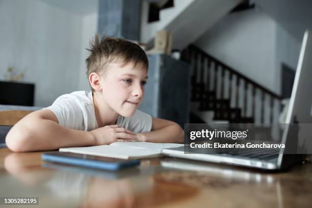 a boy of 10-11 years old in a white t-shirt sits at home at a laptop and studies online on the computer. - 10 11 years boy stock pictures, royalty-free photos & images