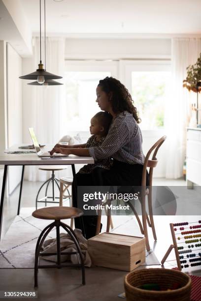 businesswoman using laptop while daughter sitting on her lap at dining table during coronavirus - leanincollection working mom stock-fotos und bilder