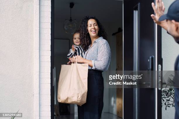 delivery man waving to smiling female customer carrying daughter while holding paper bag at doorway - food delivery stock-fotos und bilder