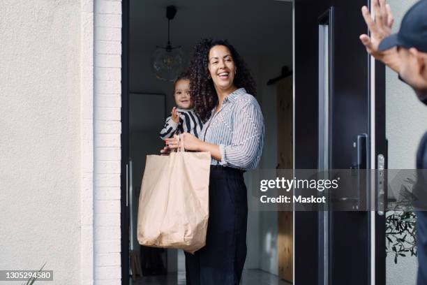 delivery man waving to smiling female customer carrying daughter while holding paper bag at doorway - baby bag bildbanksfoton och bilder