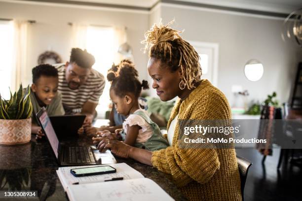 mother working from home while holding toddler, family in background - leanincollection foto e immagini stock