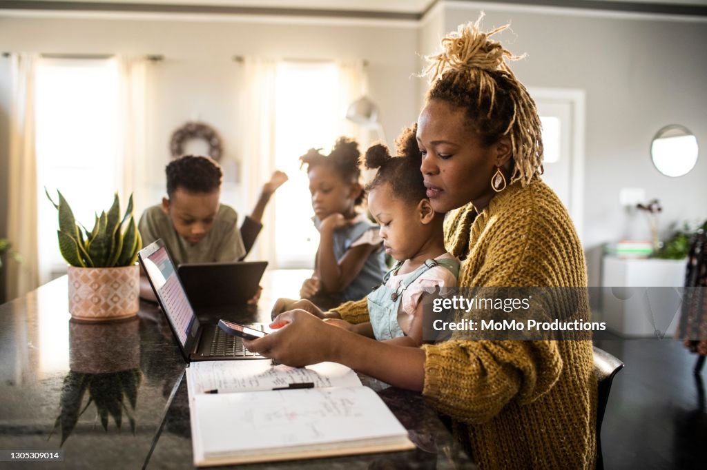 Mother working from home while holding toddler, family in background