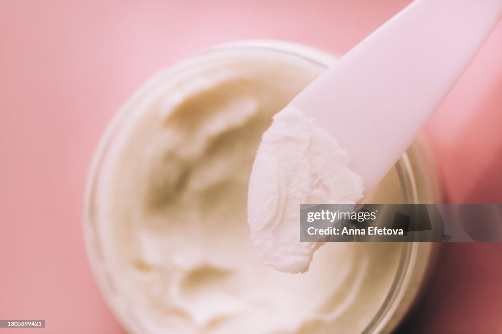 The coconut oil on spatula directly above glass jar with coconut oil on pink background. Trendy products of the year. Health and wellness concept