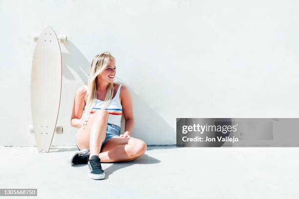 smiling blonde woman sitting on the floor with her longboard skateboard next to her leaning against the wall. - looking down photos et images de collection