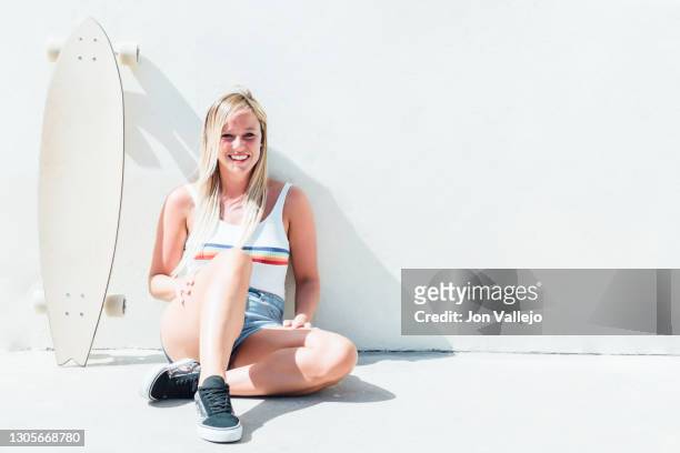 blonde woman smiling on camera and sitting on the floor with her longboard skateboard next to her leaning against the wall. - woman longboard stock-fotos und bilder