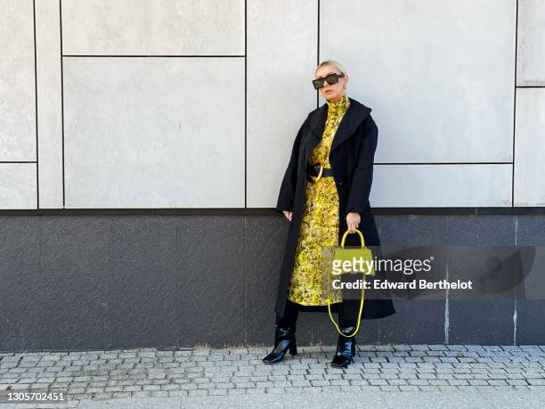 Justyna Czerniak wears sunglasses, earrings, a black long quilted/padded coat from Baum, a yellow floral print dress from Baum, a Bottega Veneta belt...