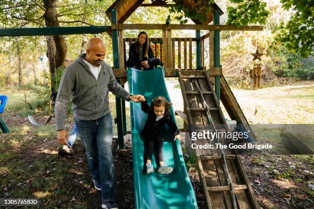 father helping daughter on slide in backyard - playground equipment happy parent stock pictures, royalty-free photos & images